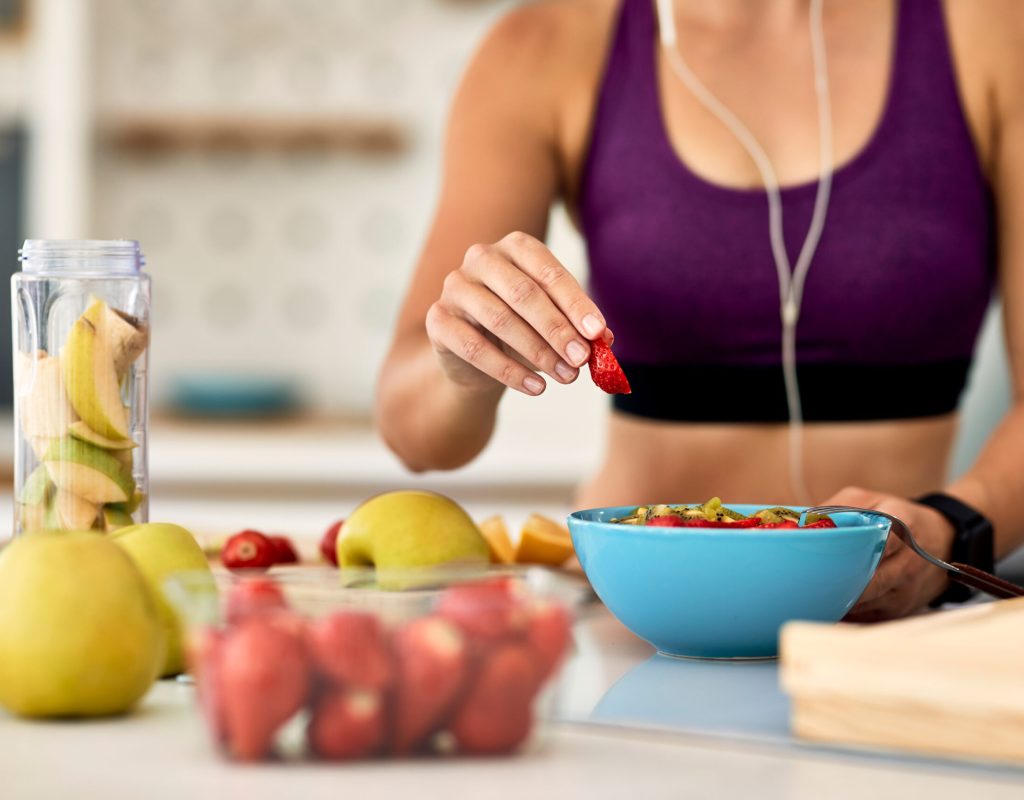 Close-up of athletic woman adding strawberries while making fruit salad in the kitchen.